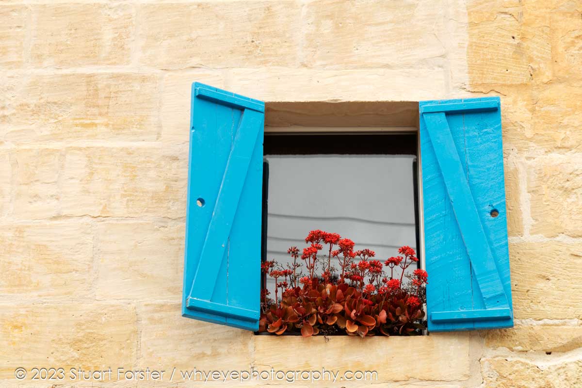 Cute window box with red flowers and blue shutters on a house in Rabat, Malta.